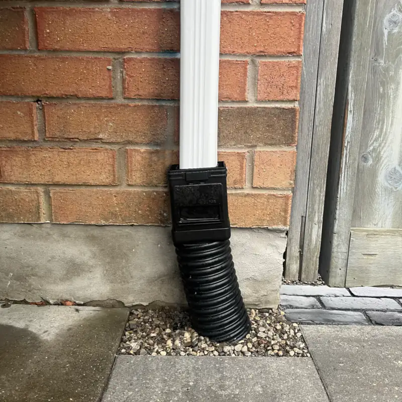 Technical drainage installation featuring a black downspout extension and drain pipe embedded in clear stone gravel